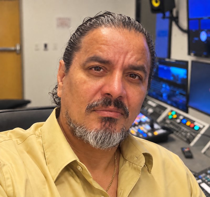 David Hernandez in a yellow shirt, seated in a broadcast control room with monitors and a switcher behind him