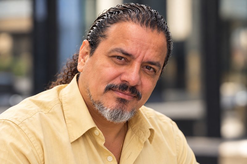 David Hernandez, headshot: long dark hair tied back, salt-and-pepper goatee, yellow button-up shirt, outdoors in Chicago